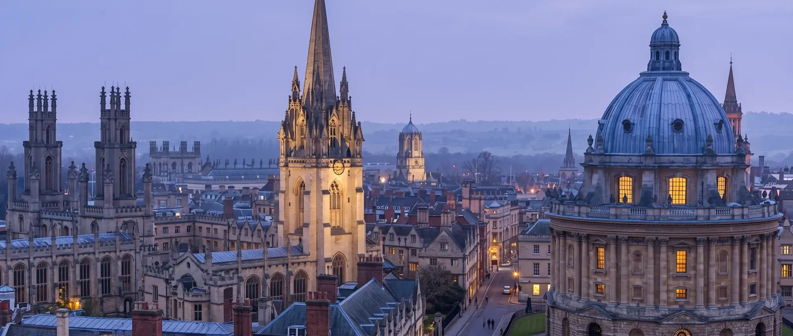 Oxford at dusk: the spire of the University Church of St Mary the Virgin, the dome of the Radcliffe Camera, and college roofs against the Oxfordshire hills.